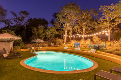 Cozy nighttime backyard: glowing kidney-shaped pool, curved terraced seating with pillows, lounge chairs, umbrellas and twinkling string lights in trees.