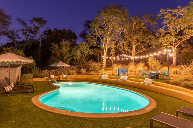 Cozy nighttime backyard: glowing kidney-shaped pool, curved terraced seating with pillows, lounge chairs, umbrellas and twinkling string lights in trees.