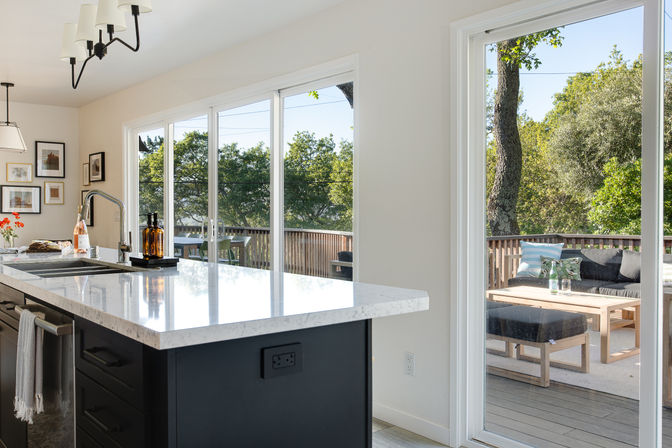 Sunlit modern kitchen with white quartz island and dark cabinets, pendant lights and sink, sliding glass doors opening to a wooden deck with cushioned outdoor seating and leafy trees.