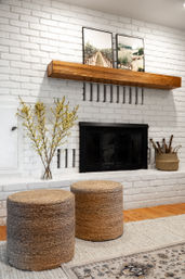 White brick fireplace with wooden mantel and landscape prints, woven jute stools, yellow branch vase and firewood basket in a cozy modern farmhouse living room