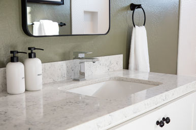 Modern bathroom vanity with white marble-look stone countertop, undermount sink and chrome faucet, two white pump dispensers, textured olive-green wall and a white hand towel on a black towel ring.