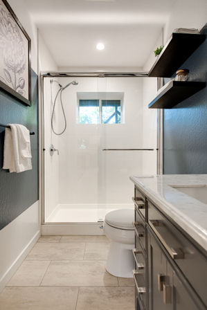Sleek modern bathroom with glass walk-in shower and handheld showerhead, small window letting in natural light, gray vanity with marble countertop, floating shelves and beige tile floor