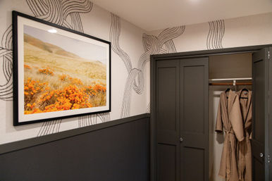 Cozy contemporary guest-room nook with framed orange poppy landscape, black-and-white wavy wallpaper, dark gray trim and an open closet showing two beige bathrobes.