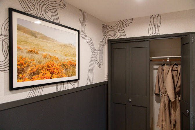 Cozy contemporary guest-room nook with framed orange poppy landscape, black-and-white wavy wallpaper, dark gray trim and an open closet showing two beige bathrobes.