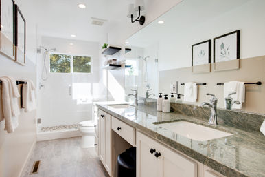 Sunlit spa-like modern bathroom with double-sink granite vanity, chrome faucets, white cabinetry, glass-enclosed walk-in shower, floating shelves, framed botanical prints and neatly folded towels.