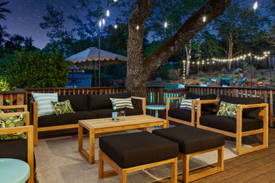 Cozy backyard patio deck at night with wooden lounge furniture and black cushions, patterned throw pillows, string lights hanging from a large tree, starry sky and a lit pool with umbrella in the background.