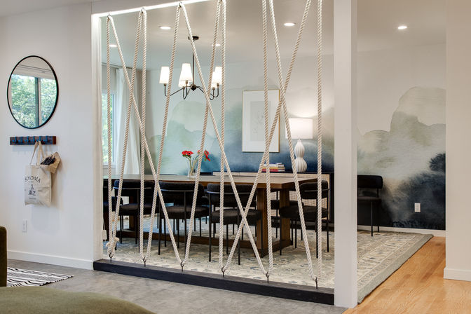 Contemporary open-plan dining room with a wooden table, black chairs, red flowers, chandelier and decorative rope partition in front of a soft watercolor mural.