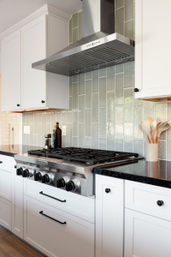 Sleek modern kitchen close-up: stainless steel six-burner gas range with vent hood, textured pale-green subway tile backsplash, white shaker cabinets, black quartz countertops, and a ceramic utensil holder with wooden spoons.