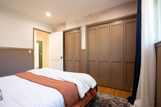 Cozy residential bedroom with brown paneled built-in closets, white bedding topped by a rust-colored quilt and cream throw, hardwood floor and patterned rug, soft natural light through white and navy curtains and an open door to a hallway.