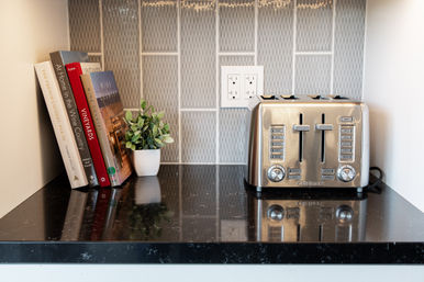 Cozy kitchen countertop nook with stainless-steel four-slice toaster, stacked cookbooks and a small potted plant on a glossy black granite surface with patterned tile backsplash and electrical outlet.