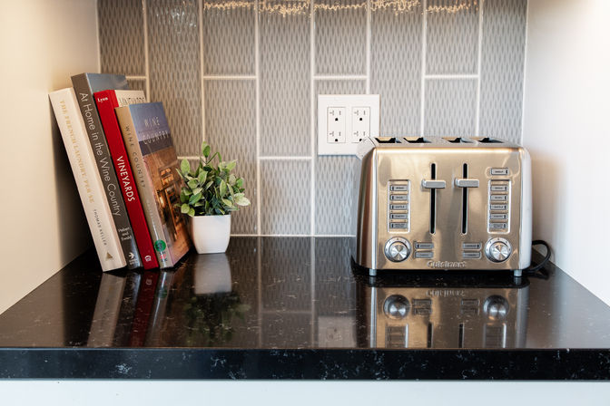Cozy kitchen countertop nook with stainless-steel four-slice toaster, stacked cookbooks and a small potted plant on a glossy black granite surface with patterned tile backsplash and electrical outlet.