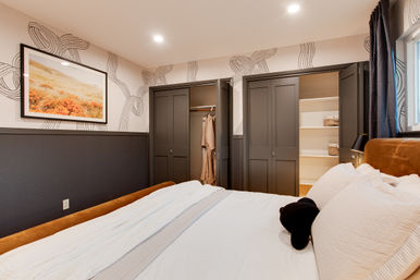 Cozy modern bedroom with white bedding, rust-colored headboard, patterned wallpaper, framed landscape art, and two open dark-gray closets showing a hanging robe and built-in shelves.