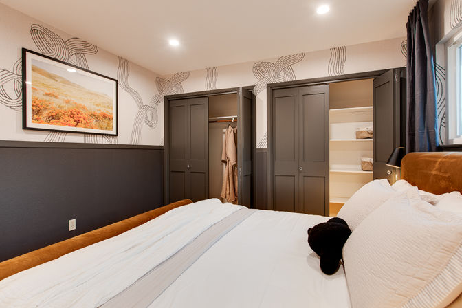 Cozy modern bedroom with white bedding, rust-colored headboard, patterned wallpaper, framed landscape art, and two open dark-gray closets showing a hanging robe and built-in shelves.