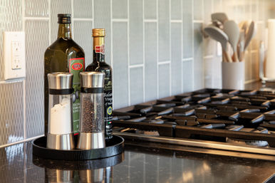 Close-up of a modern kitchen countertop showing stainless salt and pepper grinders with olive oil and balsamic bottles on a tray beside a black gas stovetop and utensil holder, ready for cooking.