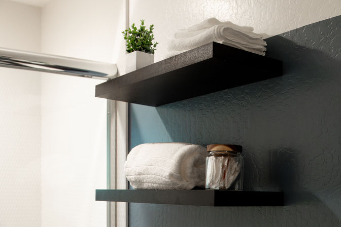 Spa-like bathroom scene with black floating shelves holding white towels, a glass jar of cotton pads, and a small potted plant against a textured blue-gray wall.