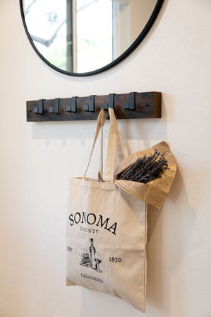 Charming canvas tote reading 'Sonoma County California' with wine illustration and dried lavender wrapped in newspaper, hanging on dark wood wall hooks under a round mirror in a bright entryway.