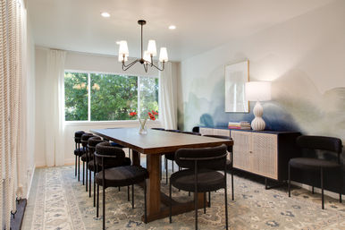 Bright, stylish dining room with a long wooden table, black upholstered chairs, large window framing green trees, blue watercolor wall mural, and rattan sideboard with lamp.
