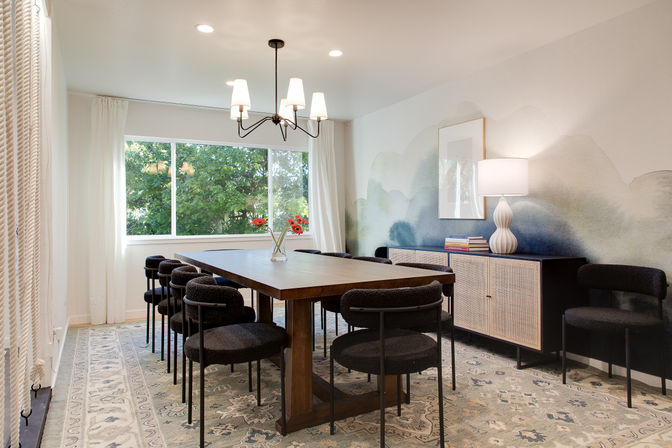 Bright, stylish dining room with a long wooden table, black upholstered chairs, large window framing green trees, blue watercolor wall mural, and rattan sideboard with lamp.