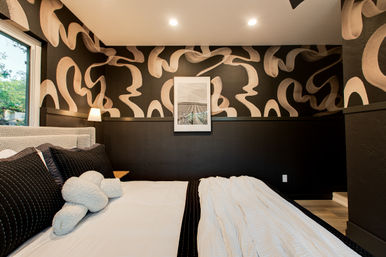 Cozy modern bedroom with bold black-and-beige abstract wallpaper, low-profile bed dressed in black pillows and white bedding, bedside lamp, framed wall art, and a window view of greenery.
