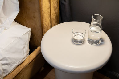 Round white bedside table with a glass and carafe of water next to a beige upholstered bed with white linens and gold curtain.