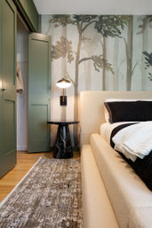 Cozy modern bedroom corner with a cream upholstered bed, black-striped bedding, black marble round nightstand and brass wall sconce, green paneled closet doors, watercolor forest mural wallpaper, hardwood floor and patterned rug.