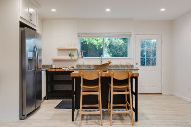 Bright modern home kitchen with granite-topped island and two wooden bar stools, stainless steel refrigerator, open shelving, window looking out to leafy backyard and white back door.