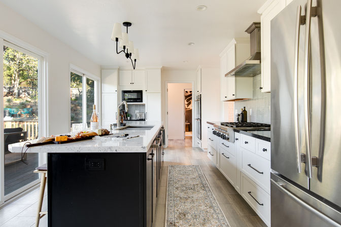 Sunny modern open-concept kitchen with a large marble-topped island, white shaker cabinets, stainless steel refrigerator and range, pendant lights, sliding glass doors to a deck, and a patterned runner rug.