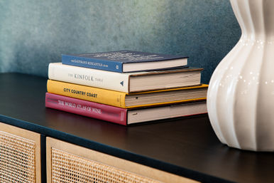 Stylish stack of hardcover lifestyle books on a black sideboard beside a white ceramic vase, woven cane cabinet doors and textured teal wall — modern home decor vignette.