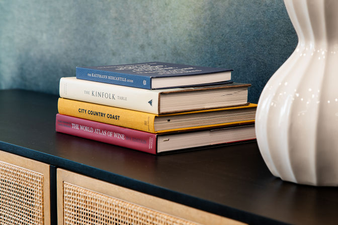 Stylish stack of hardcover lifestyle books on a black sideboard beside a white ceramic vase, woven cane cabinet doors and textured teal wall — modern home decor vignette.