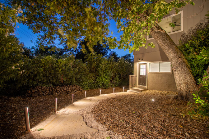 Cozy twilight residential backyard with a curved stone pathway lined by modern LED bollard lights leading to a house entrance under a large leaning oak tree, surrounded by shrubs and mulch.