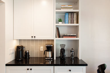 Modern home coffee station on black granite countertop under white cabinets — drip coffee maker, bean grinder and French press with cookbooks and a small potted plant on open shelves.