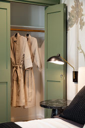 Cozy bedroom scene with open sage-green closet doors showing two beige waffle-knit bathrobes on wooden hangers, next to a black-and-gold wall lamp and round black marble bedside table.