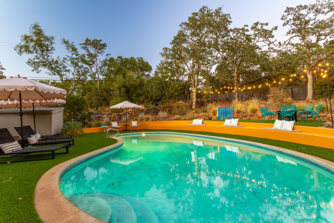 Inviting backyard kidney-shaped pool with turquoise water and curved steps, surrounded by green turf, scalloped parasols and black lounge chairs, a bright yellow terraced wall with pillows, teal patio seating, and string lights strung through oak trees.