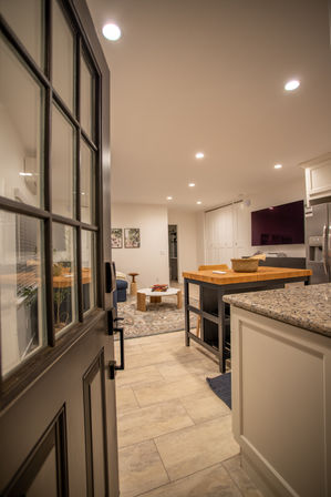 Open black front door opening to a cozy modern open-concept kitchen and living room with a wood-topped island, granite countertop, tiled floor, recessed ceiling lights, rug, coffee table and wall-mounted TV.