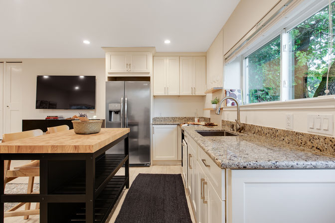 Bright modern open-concept kitchen with granite countertops, stainless steel fridge, butcher-block island and large window overlooking trees.