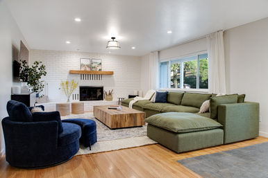 Cozy modern living room with oversized green sectional chaise, plush navy swivel chairs, white brick fireplace, rustic wooden coffee table and a large window letting in natural light
