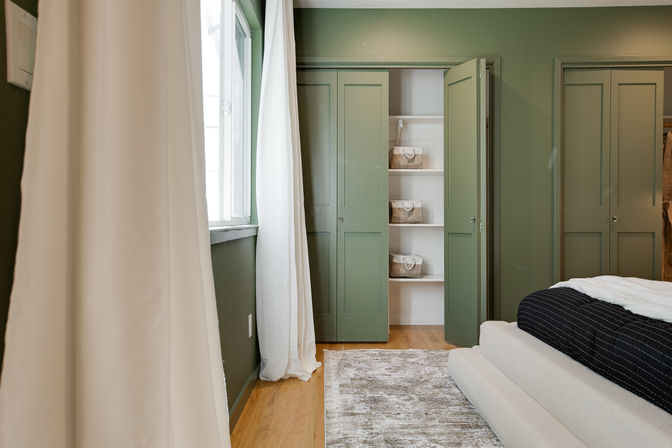 Cozy green bedroom interior with white curtains and natural light, wood floor, neutral rug, low platform bed with black-and-white bedding, and open green closet showing white shelves with woven storage baskets.