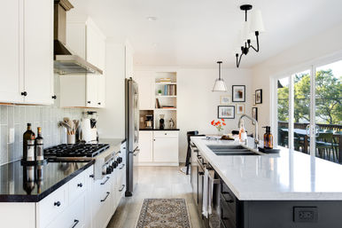 Bright open-concept modern kitchen with white cabinets, stainless steel range and fridge, large white quartz island with sink and bar seating, pendant lights, rug runner, vase of orange flowers on the counter, and sliding glass doors opening to a sunny deck and trees.