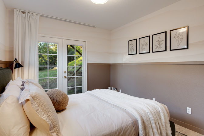 Cozy neutral-toned bedroom with a made bed, textured pillows and throw, framed botanical prints on the wall, and glass French doors opening to a leafy backyard.