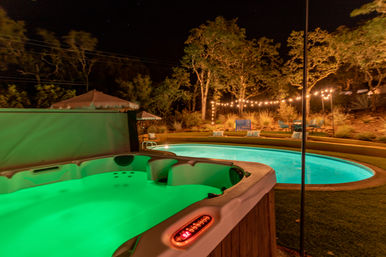 Nighttime backyard oasis featuring a green-lit hot tub in the foreground and a glowing kidney-shaped pool with string lights, patio seating and trees in a landscaped yard.