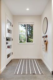 Sunlit modern entryway with white glass-paneled front door, window overlooking leafy yard, round wall mirror, wooden hooks holding a canvas tote, and a black-and-white striped rug on gray tile flooring.