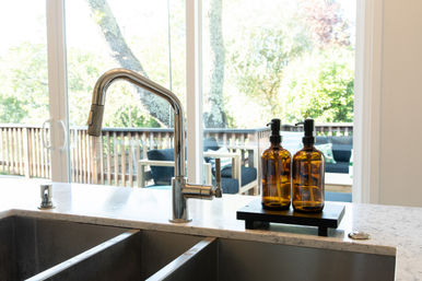 Modern residential kitchen with a stainless steel faucet and double sink on a marble countertop, amber pump soap bottles on a small tray, and sliding glass doors opening to a sunny wooden deck and backyard.