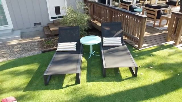 Two black outdoor lounge chairs with striped pillows flanking a small round white table on bright green artificial grass next to a wooden deck in a sunny backyard patio.