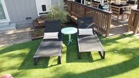 Two black outdoor lounge chairs with striped pillows flanking a small round white table on bright green artificial grass next to a wooden deck in a sunny backyard patio.