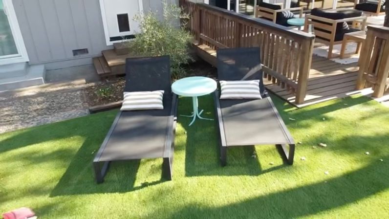 Two black outdoor lounge chairs with striped pillows flanking a small round white table on bright green artificial grass next to a wooden deck in a sunny backyard patio.