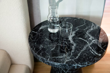 Sleek round black marble side table with white veining holding a glass carafe half-filled with water and an empty tumbler beside a beige sofa in a modern living room