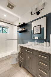 Modern residential bathroom with navy accent wall, gray vanity and marble countertop, glass shower, floating shelves, mirror and beige tile floor.