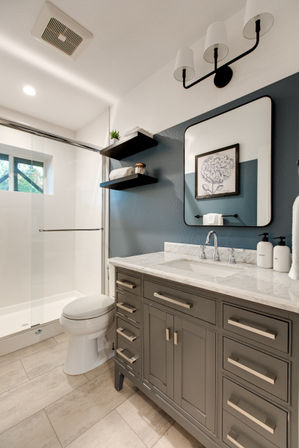 Modern residential bathroom with navy accent wall, gray vanity and marble countertop, glass shower, floating shelves, mirror and beige tile floor.