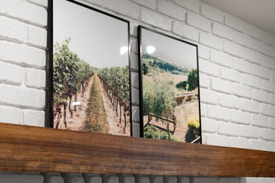 Two black-framed landscape prints—vineyard rows and sunlit countryside—leaning on a wooden mantel against a white painted brick wall.