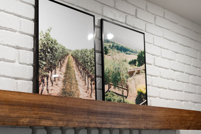 Two black-framed landscape prints—vineyard rows and sunlit countryside—leaning on a wooden mantel against a white painted brick wall.